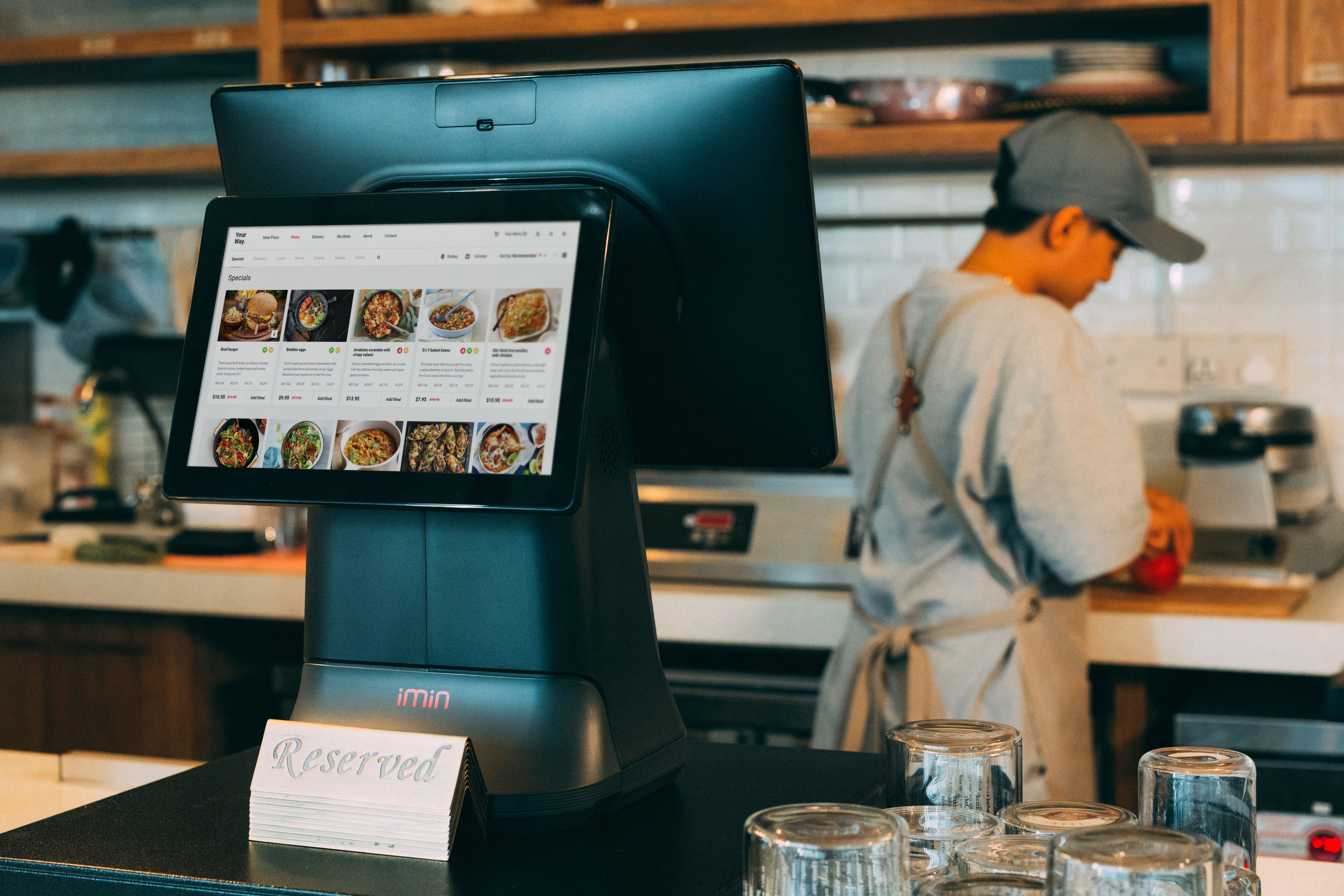 A chef working beside a touchscreen digital ordering system in a restaurant kitchen.
